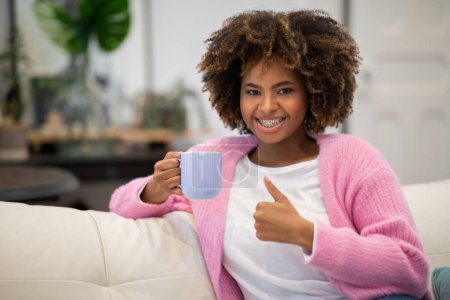 Foto de Happy cheerful pretty curly young black woman in casual enjoying fresh aromatic coffee, holding mug and showing thumb up at camera, lady drinking herbal tea in the morning at home, copy space - Imagen libre de derechos