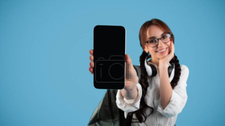 Photo for Glad caucasian teenage girl with pigtails in glasses, student shows smartphone with blank screen, isolated on blue background, panorama, studio. Website for study, news, app for education, knowledge - Royalty Free Image