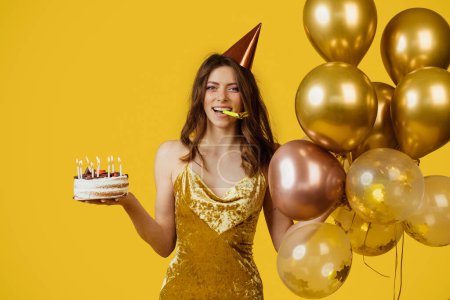 Téléchargez les photos : Femme joyeuse en robe et chapeau d'anniversaire, tenant gâteau avec des bougies et des bouquets de ballons d'air, soufflant dans la pipe, célébrant fête d'anniversaire de vacances sur fond jaune studio. - en image libre de droit