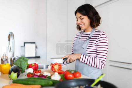 Téléchargez les photos : Happy beautiful young brunette middle eastern lady wearing grey apron enjoying cooking at home, woman cutting vegetables and smiling, preparing healthy meal, using only organic veggies, copy space - en image libre de droit