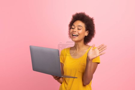 Téléchargez les photos : Laughing young african american curly woman in yellow t-shirt waving hand at laptop webcam, say hello isolated on pink background, studio. Greeting, video call, meeting remotely, chat and lesson - en image libre de droit