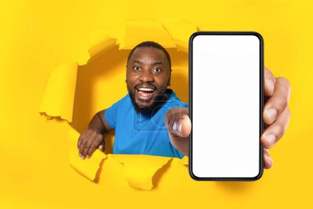 Téléchargez les photos : Happy african american man holding big cellphone with blank screen in hand, showing gadget with mockup for app or website through torn yellow paper hole, collage - en image libre de droit