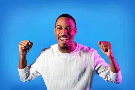 Téléchargez les photos : Cool happy handsome young african american man in casual outfit gesturing on colorful neon studio background, emotional guy clenching fists in neon light, copy space for advertisement - en image libre de droit