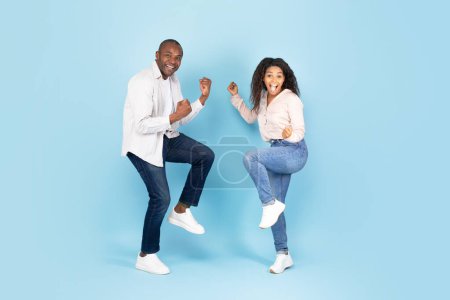 Foto de Wow, yes. Emotional black man and woman shaking clenched fists, looking at camera with excitement, celebrating win on blue studio wall, full body length banner - Imagen libre de derechos
