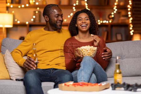 Téléchargez les photos : Happy beautiful adult black couple spending weekend together at home, loving man and woman sitting on couch, bonding, drinking beer, eating popcorn and pizza, watching movie, laughing, copy space - en image libre de droit