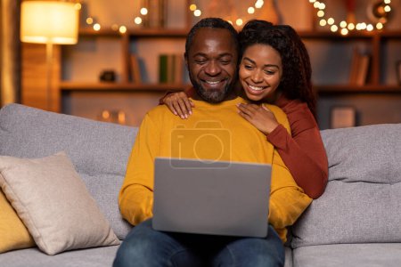 Téléchargez les photos : Happy cheerful loving black spouses middle aged man and millennial woman shopping on Internet while resting at home, looking at laptop screen and smiling, copy space. Retail, e-commerce concept - en image libre de droit