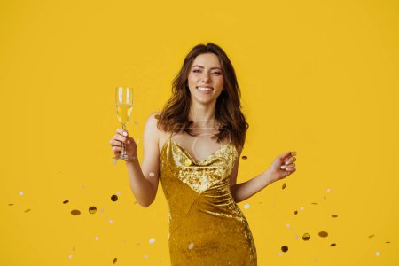 Téléchargez les photos : Posh party. Jeune femme glamour en robe de soirée festive debout avec une coupe de champagne sous les confettis tombants et souriant à la caméra, fond studio jaune - en image libre de droit