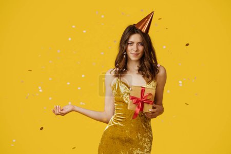 Téléchargez les photos : Heureuse dame tenant des boîtes-cadeaux enveloppées, portant une robe de fête et un chapeau d'anniversaire sur fond de studio jaune avec des confettis tombants. Belle femme faisant la fête du jour-B - en image libre de droit