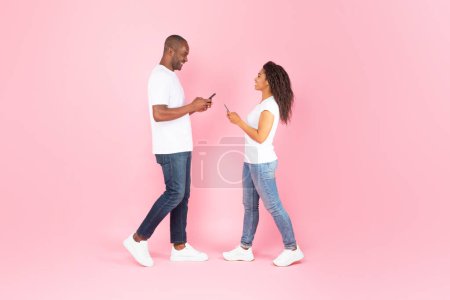 Photo for Black middle aged man and young woman using smartphones, standing over pink studio background, full length shot, side view. New mobile application ad concept - Royalty Free Image
