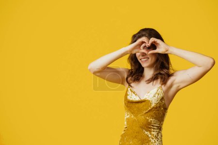 Foto de Mujer bonita sonriente mostrando el signo del corazón con las manos cerca de los ojos, posando en vestido sobre fondo amarillo, tiro al estudio, espacio libre. Signo de amor, gratitud, romance y agradecimiento - Imagen libre de derechos