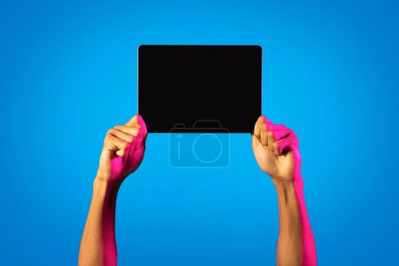 Téléchargez les photos : Online offer, mobile app. Cropped of african american man hands hand holding horizontal digital tablet with blank screen on blue studio background in neon light, mockup for advertisement, copy space - en image libre de droit