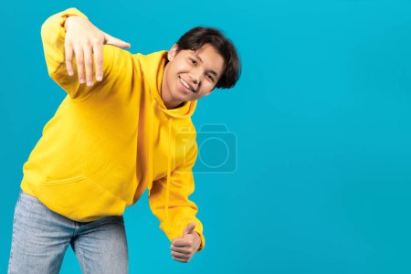 Téléchargez les photos : Cheerful Japanese Teenager Boy Posing Offering Hand For Handshake And Gesturing Thumbs Up Smiling To Camera Standing Near Copy Space Advertising Great Offer On Blue Studio Background - en image libre de droit