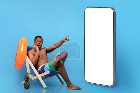 Foto de Millennial black guy in swimwear sitting in lounge chair, pointing at phone with blank screen in surprise on blue studio background, mockup. Summertime sale or discount, seasonal promo, collage - Imagen libre de derechos