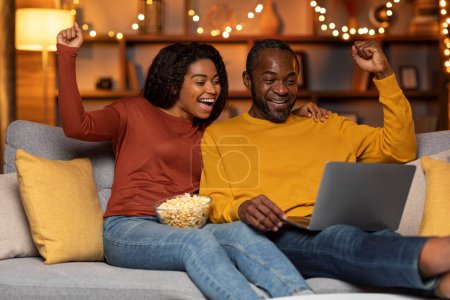 Foto de Emotional happy african american family husband and wife sitting on couch with laptop and bowl full of delicious popcorn, celebrating success, trading online, cozy home interior - Imagen libre de derechos