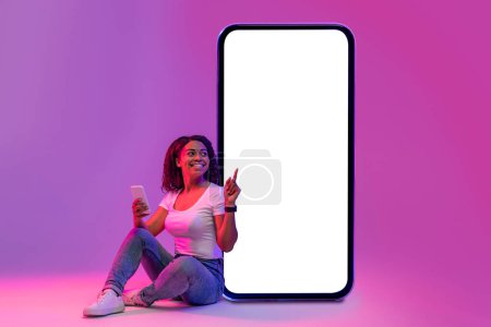 Téléchargez les photos : Smiling Black Woman With Smartphone In Hand Sitting In Neon Light And Pointing At Huge Blank Telephone Screen, Happy African Female Recommending New Mobile Application, Creative Collage, Mockup - en image libre de droit