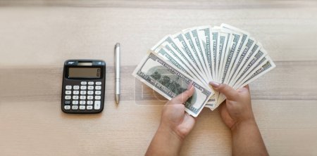 Photo for Female hands holding bunch of cash dollars, pen and calculator on wooden table. Unrecognizable dark-skinned woman working on budget, counting monthly expenses, top view, cropped. Finance, loan concept - Royalty Free Image