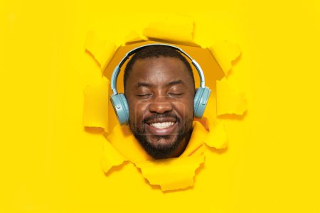 Téléchargez les photos : Homme afro-américain détendu profitant de la musique dans des écouteurs avec les yeux fermés, posant et souriant à travers trou déchiré de fond de papier jaune - en image libre de droit