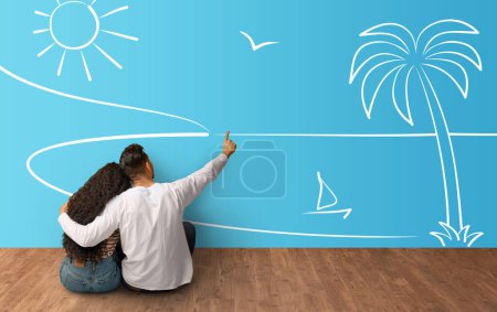 Photo for Back view of middle eastern young loving couple sitting on floor, embracing and pointing at seaside sketch on blue, dreaming about vacation or house next to sea, collage - Royalty Free Image
