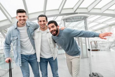 Téléchargez les photos : Voyagez avec des amis. Trois touristes joyeux gars étreignant posant avec des valises et s'amusant dans l'aéroport moderne à l'intérieur, souriant à la caméra. Groupe d'hommes voyageant à l'étranger ensemble. Concept de tourisme - en image libre de droit