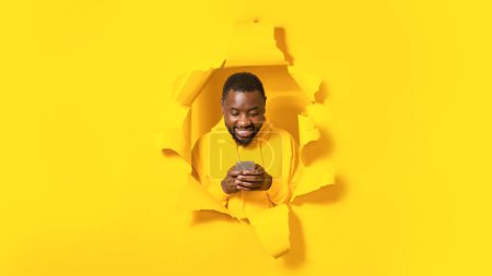 Téléchargez les photos : Happy african american man using smartphone and browsing internet posing in hole in torn yellow paper background, panorama. Great mobile application concept. Collage - en image libre de droit