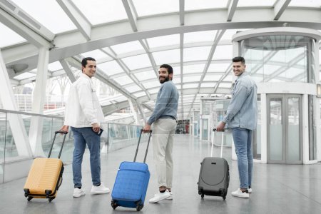Téléchargez les photos : Group Of Male Tourists Walking With Suitcases Smiling To Camera Posing In Airport Terminal Indoors. Three Friends Men Traveling Together On Vacation. Tourism And Transportation Concept. Full Length - en image libre de droit