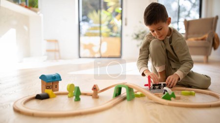 Foto de Glad european small kid in pajamas plays with toys, train, cars and wooden road, enjoy spare time, sits on floor in bright living room interior, panorama. Fun alone, fantasy at home and kindergarten - Imagen libre de derechos