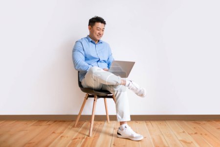 Téléchargez les photos : Positive middle aged asian man using laptop, surfing internet while sitting in chair against white studio wall, free space, full length. E-learning, online lectures and courses - en image libre de droit