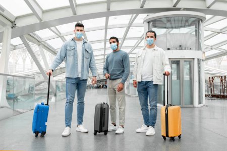 Téléchargez les photos : Bon voyage. Trois touristes les gars portant des masques de visage posant debout avec des valises dans l'aéroport moderne à l'intérieur. Groupe de voyageurs masculins voyageant à l'étranger pendant la pandémie de Covid-19 - en image libre de droit