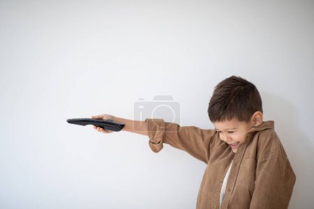 Photo for Cheerful european small kid laughs uses remote control, switches channel of TV, shoots at imaginary gun and plays on white wall background, studio, empty space. Fun at home with technology, childhood - Royalty Free Image
