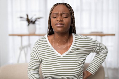 Photo for Back pain, causes and treatment concept. Upset young long-haired black woman in pain suffering from backache, touching lower back while sitting on sofa at home, closeup, copy space - Royalty Free Image