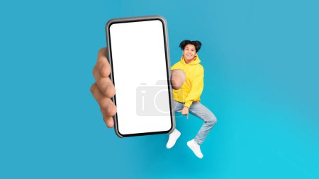 Foto de Cheerful Asian Teenager Boy Showing Large Cellphone With Empty Screen Jumping On Blue Studio Background. Teen Recommending New Mobile App Or Great Offer Holding Phone. Panorama, Mockup - Imagen libre de derechos