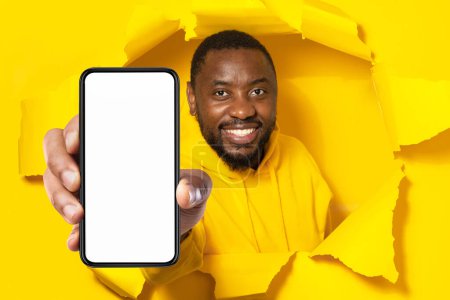 Foto de Recomendación de la aplicación. Hombre negro feliz mostrando un gran teléfono inteligente con pantalla en blanco a través de un agujero de papel amarillo roto. Gadget con espacio libre vacío para maqueta, bandera - Imagen libre de derechos