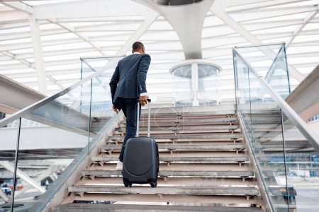 Foto de Black Businessman With Suitcase Walking Up Stairs At Airport Terminal, Rear View Of Young African American Man Wearing Suit Carrying Luggage While Going To Flight Departure Gate, Copy Space - Imagen libre de derechos