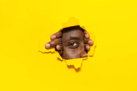 Foto de Te estoy observando. Hombre afroamericano mirando a la cámara a través de un agujero en un fondo de papel amarillo roto, espiando y espiando, de cerca. Concepto de curiosidad - Imagen libre de derechos