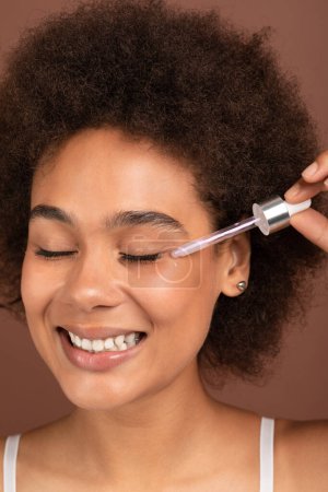Foto de Smiling young african american curly lady with perfect skin apply serum on her face, isolated on brown background, studio, close up. Beauty care, cosmetics and spa anti-aging treatments, cosmetology - Imagen libre de derechos