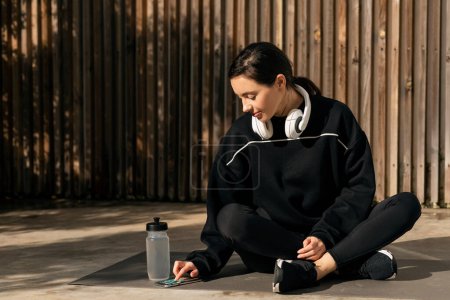 Téléchargez les photos : Glad caucasian millennial lady in sportswear and headphones rest, sit on floor, typing on smartphone in street with bottle of water. Sports blog, app for body care and weight loss, fitness with device - en image libre de droit