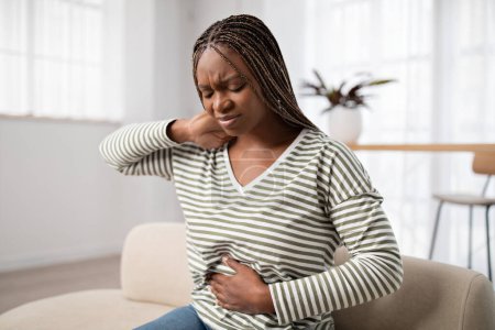 Photo for Unhappy young black woman wearing casual clothes suffering from menstrual cramps, feeling sick to her stomach, holding belly and touching neck, pain during period, sitting on couch at home - Royalty Free Image