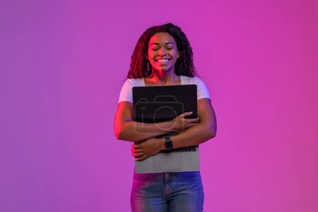 Téléchargez les photos : Happy Black Woman Embracing Laptop Computer With Blank Screen While Standing In Neon light Over Purple Background, Cheerful African American Lady Hugging Pc, Enjoying Modern Technologies, Copy Space - en image libre de droit