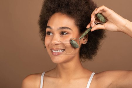 Foto de Glad pretty young black curly female with perfect skin applies powder or blush with brush on face, isolated on brown background, studio. Ad and offer of cosmetics, nude make-up lesson, beauty care - Imagen libre de derechos