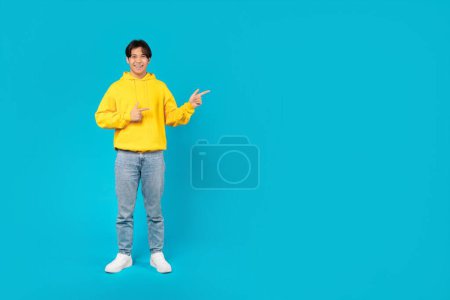 Photo for Positive Asian Teen Boy Pointing Fingers Aside Standing Near Empty Space For Text Recommending Great Offer Posing Over Blue Studio Background. Look There Concept. Full Length Shot - Royalty Free Image