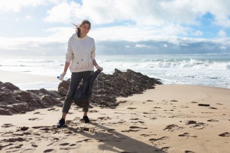 Téléchargez les photos : Journée mondiale de l'environnement. Volontaire femme debout sur la plage avec sac, ramassage des ordures et nettoyage de la zone côtière, espace de copie, pleine longueur - en image libre de droit
