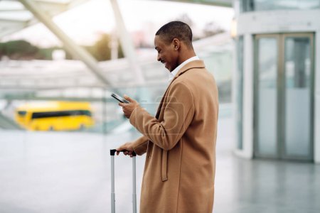 Téléchargez les photos : Smiling Black Man Standing With Suitcase Outdoors And Using Smartphone At Airport, Happy Young African American Male Booking Transfer Online Or Browsing Taxi App After Arrival To New City, Copy Space - en image libre de droit