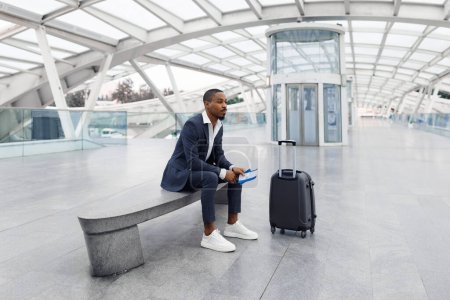 Téléchargez les photos : Portrait Of Pensive Young Black Businessman Sitting On Bench At Airport Terminal, African American Male Wearing Suit Holding Passport With Tickets, Waiting For Flight Boarding, Copy Space - en image libre de droit