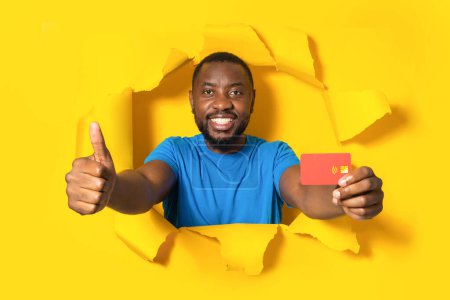 Foto de Excited african american man holding credit card and showing thumb up posing through torn yellow paper background. Male recommending bank services - Imagen libre de derechos