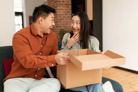 Photo for Surprised korean couple unpacking box parcel after successful shopping, sitting on sofa in living room at home, copy space. Asian spouses opening cardboard package, receiving order from online store - Royalty Free Image