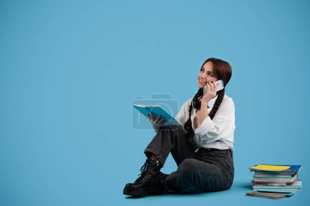 Téléchargez les photos : Happy caucasian teenager girl with pigtails, student sits on floor, reads books, calls on phone, looks at empty space, isolated on blue background, studio. Communication, learn, knowledge, education - en image libre de droit