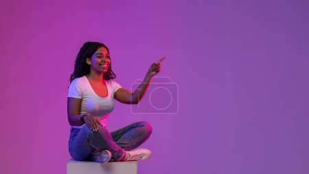 Photo for Beautiful Black Woman Sitting On Big Cube In Neon Light And Pointing Aside At Copy Spacen Over Gradient Purple Background, Happy African American Lady Showing Free Place For Advertisement, Panorama - Royalty Free Image