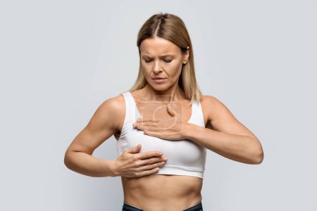 Téléchargez les photos : Breast screening. Anxious middle aged blonde woman wearing white top examining her breasts, grey studio background, cancer prevention concept, copy space - en image libre de droit