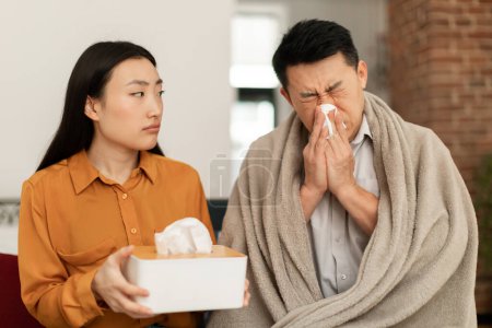 Foto de Sick asian man wrapped in blanket sitting on sofa and sneezing nose, young wife treating her ill middle aged husband, taking care and giving napkins, seasonal viruses and flu concept - Imagen libre de derechos