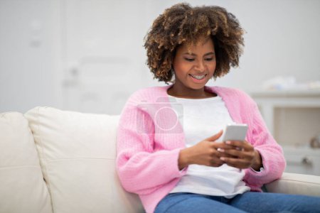 Téléchargez les photos : Happy smiling beautiful black lady with bushy hair sitting on couch with smartphone, reading blog, scrolling on social media, home interior, copy space for ad, nice mobile app concept - en image libre de droit